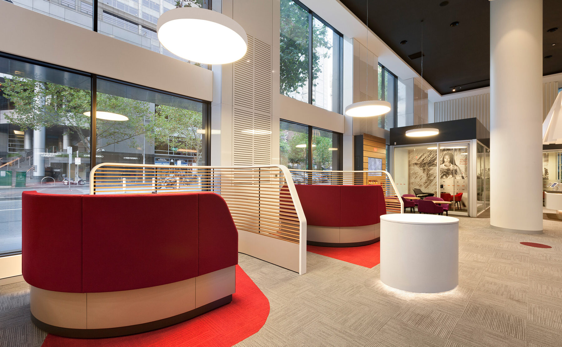 Bank customer lounge, red upholstered booths, curved partition screen, halo pendant lighting, circular plinth, double height curtain wall, commercial carpet tile.
